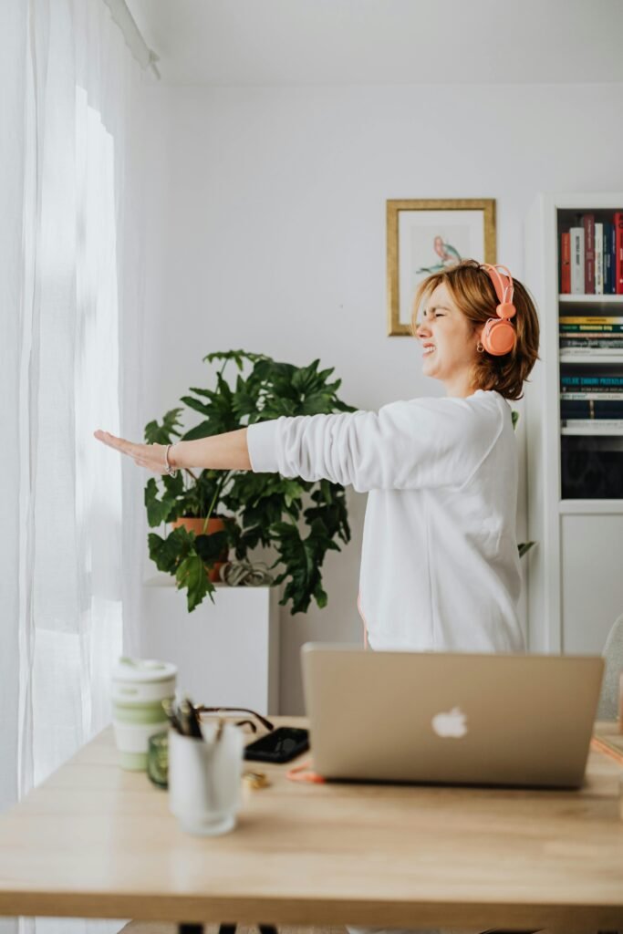 Woman stretching in a bright home office wearing headphones, with a laptop on the desk.